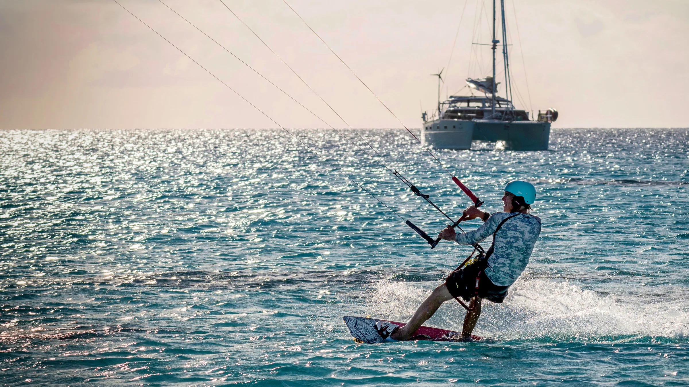 Kitesurfers crossing bright water with catamarans at anchor in the distance